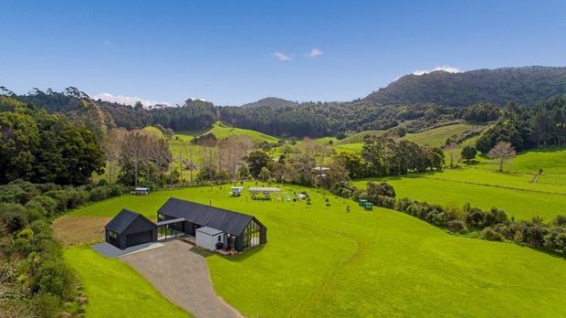 Aerial view of a contemporary rural property set in expansive vivid green farmland, with a dark-clad low-rise building, gravel driveway, surrounding trees, rolling hills and forested mountains in the background under a clear blue sky.