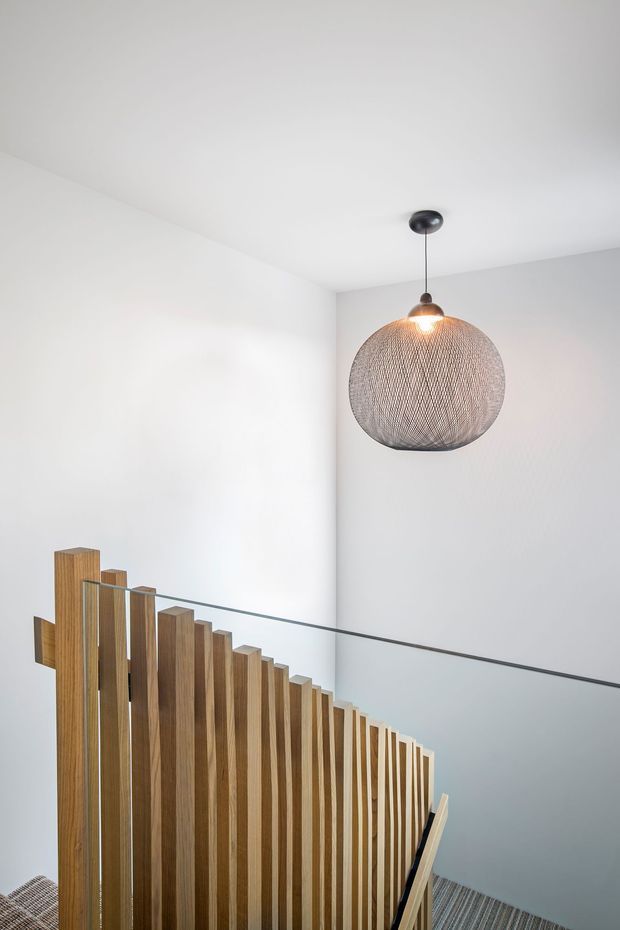 A minimalist interior stairway viewed from the upper landing, featuring a sculptural open timber balustrade with closely spaced vertical slats, a clear glass guardrail, white walls and ceiling, grey carpeted stairs, and a large woven spherical pendant light.
