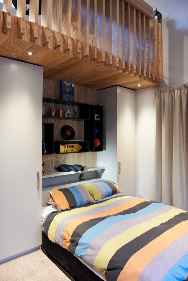 Compact, colourful bedroom with a double bed dressed in bold horizontal striped bedding. The room features extensive warm timber wall panelling and a dramatic timber overhead loft or mezzanine balustrade with vertical slats. Built-in white wardrobes flank the bed, while a recessed entertainment and display unit includes dark shelving, a television, records and decorative objects. Full-height curtains cover the window, and recessed ceiling lights provide warm illumination.