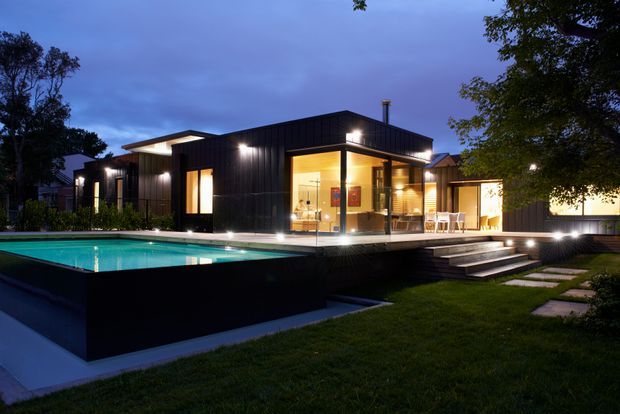 Twilight exterior view of a contemporary single-storey home with dark vertical cladding, extensive warm-lit glazing, a raised swimming pool, timber deck, broad steps, and landscaped lawn. The house has a low, flat roof form and strong indoor-outdoor connection.
