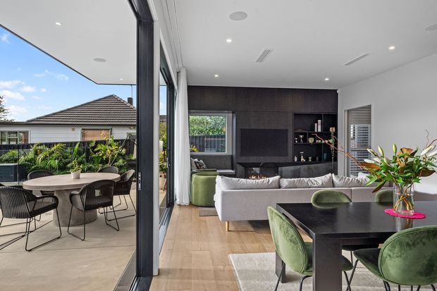 Contemporary open-plan living and dining area opening onto a covered outdoor terrace. The interior features pale timber flooring, a large light sectional sofa, dark built-in media cabinetry with a fireplace, a black dining table, and green upholstered dining chairs. Full-height glazing connects the room to the terrace, which has an outdoor dining setting and views toward a suburban residence. The palette is refined and neutral with deep charcoal joinery and green accents.