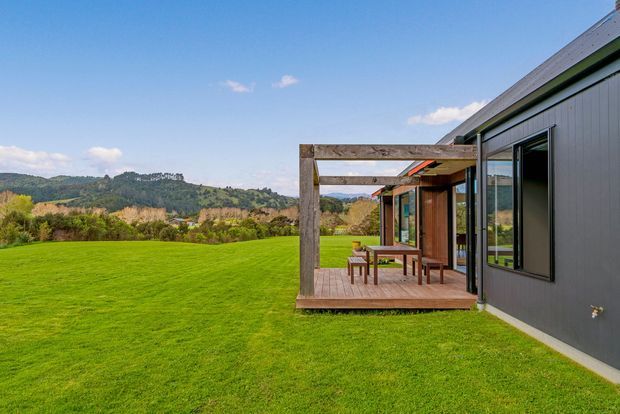 Exterior view of a contemporary dark-clad building opening onto a timber deck with a simple pergola and outdoor dining setting. The building sits beside a broad manicured lawn, with rolling forested hills visible in the background under a clear sky.
