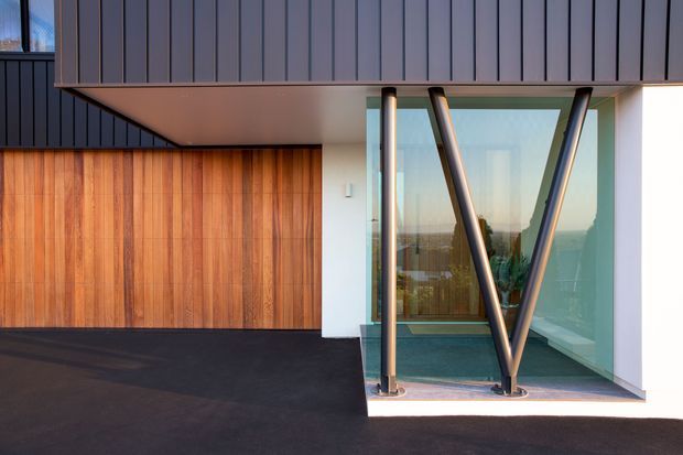 A contemporary residential exterior featuring dark vertical metal cladding, a warm timber-clad garage door, and a dramatic glazed entry or corner window framed by angled dark structural supports. The building has crisp white rendered walls, a projecting flat roof canopy, and a dark paved driveway. Reflections reveal interior greenery and distant landscape beyond the glass.