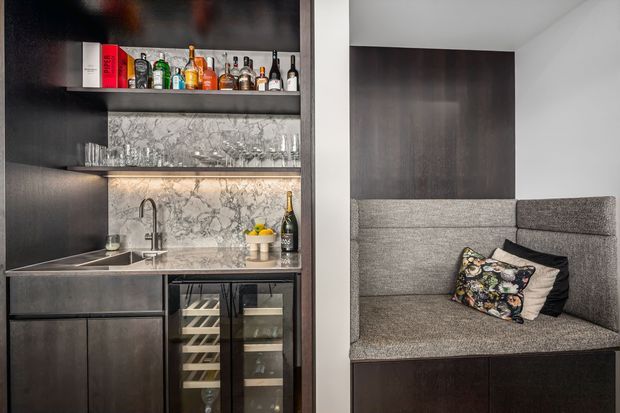 A compact, sophisticated built-in home bar and kitchenette beside a fully upholstered grey banquette. The bar features dark timber cabinetry, open shelving with bottles and glassware, a marble-look splashback and benchtop, integrated sink and under-counter wine storage. The adjacent seating nook has a high upholstered back and colourful decorative cushions.