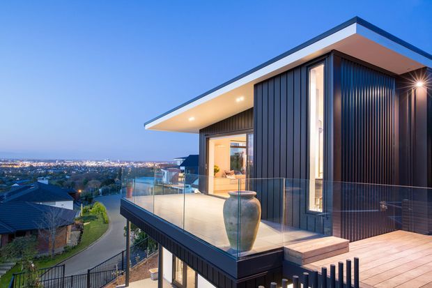 A contemporary multi-level home viewed from an elevated exterior terrace at dusk, with a dramatic flat roof, deep projecting eaves, dark vertical cladding, extensive glazing, and clear glass balustrades. The terrace overlooks a suburban cityscape and appears to include outdoor seating and a large decorative planter. Warm interior lighting contrasts with the dark facade and blue evening sky.