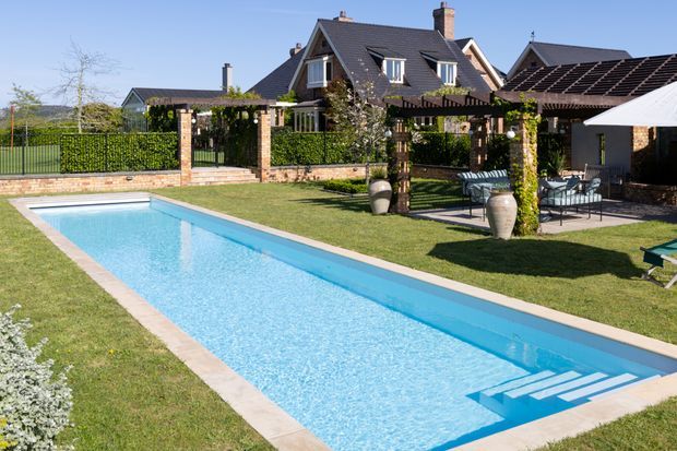 Sunny landscaped backyard with a long rectangular swimming pool, pale paved coping, manicured lawn, and a covered outdoor entertaining area with pergola, dining furniture, planters, and a large umbrella. A substantial pitched-roof brick-and-render house and garden fencing are visible in the background.