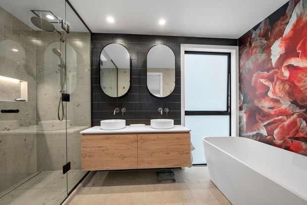 A contemporary bathroom featuring a walk-in glass shower with large-format grey stone-look wall tiles, a floating timber vanity with two white basins, black vertical wall tiles, rounded mirrors, a freestanding white bathtub, and a bold red and charcoal floral feature wall. The room has a clean, high-contrast, refined aesthetic with recessed ceiling lighting and frosted glazing.