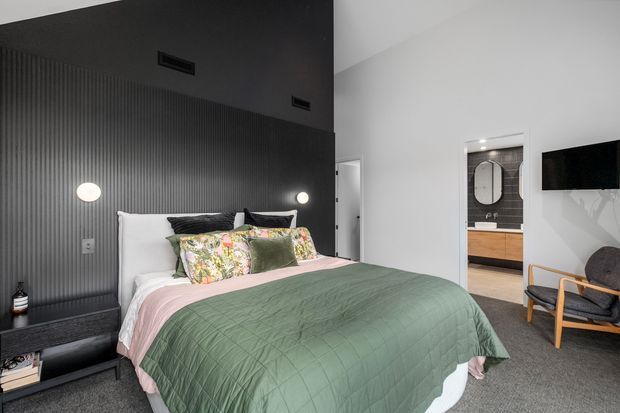 Contemporary bedroom with a large bed dressed in a muted green quilt and blush pink bedding, layered floral and neutral cushions, and a dark vertically ribbed feature wall. Wall-mounted globe lights flank the bed, while a floating dark bedside shelf, wall-mounted television, upholstered timber-framed armchair, and adjoining vanity area complete the room.