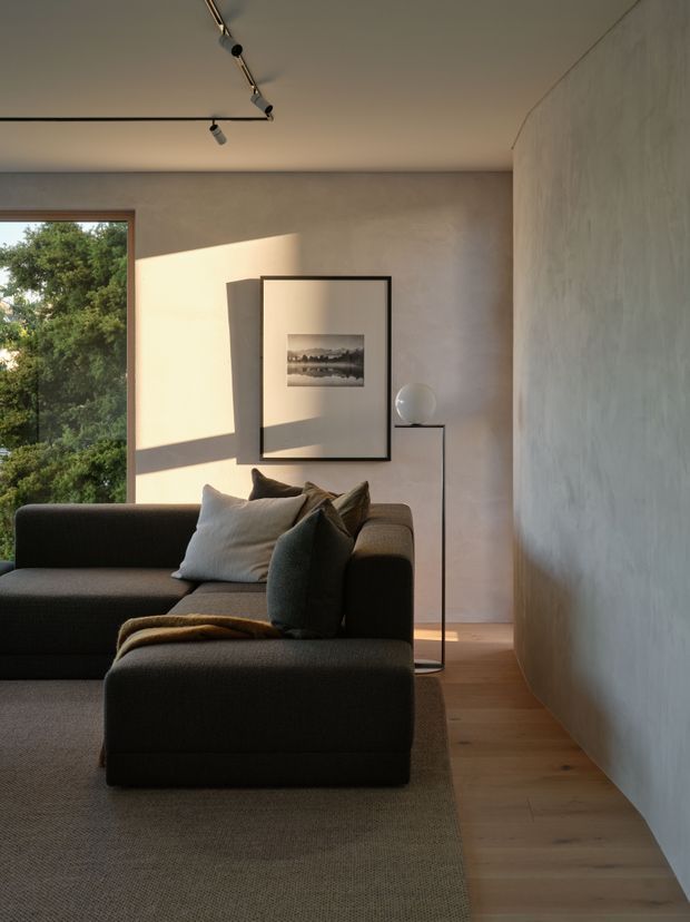 A softly lit contemporary living room with a large dark sectional sofa, neutral cushions and throw, pale textured plaster walls, light timber flooring, and a simple black-framed artwork with a slender floor lamp. Full-height glazing at left brings in views of leafy greenery, while track lighting and a large neutral rug complete the calm, minimalist interior.