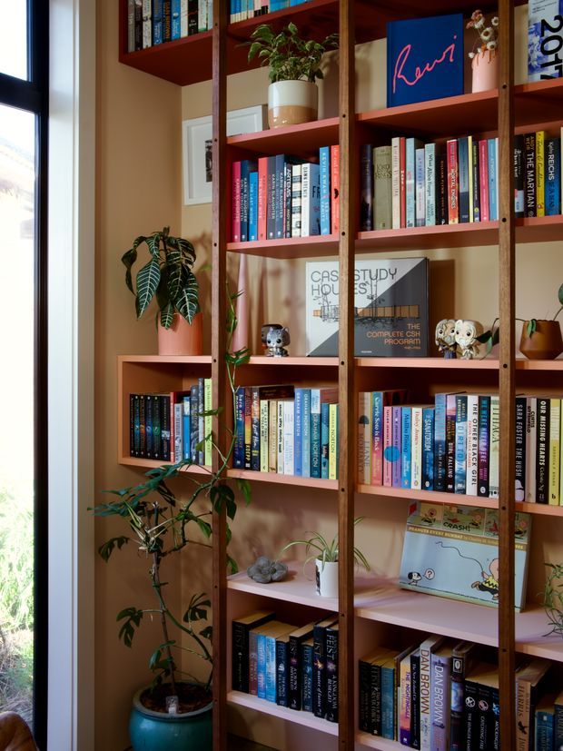 A tall custom open bookshelf fills the frame beside a large window, with warm wood uprights, muted pink painted shelves and a tan wall. The shelving is densely arranged with books, framed artwork, ceramic objects, and several potted plants, creating a colourful, personal library nook with abundant natural light.