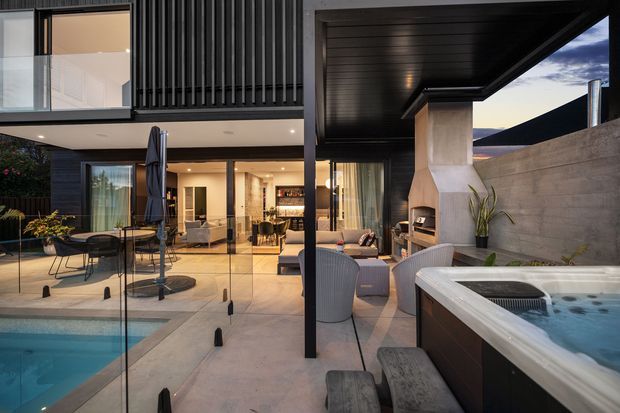 Contemporary multi-level home viewed from a covered outdoor entertaining area, with a swimming pool in the foreground, a spa, glass balustrades, an outdoor fireplace, and a glazed living area opening onto the terrace. The architecture features strong dark vertical cladding, a deep black soffit, concrete walls, and clean rectilinear forms.