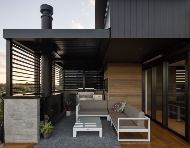 A sophisticated covered outdoor entertaining area beside a dark-clad contemporary home. The space includes a built-in outdoor kitchen and barbecue, a lounge setting with a sectional sofa and coffee table, timber decking, concrete and dark metal finishes, horizontal privacy screens, and sliding glass doors opening to the interior. A cylindrical flue and roof structure emphasize the sheltered architectural form, with a landscape view beyond the screens.
