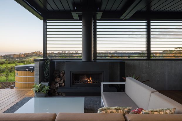 Covered outdoor living area with a built-in fireplace, timber decking, a large sectional sofa, coffee table, horizontal louvred screening, and a countryside view beyond. The space has a dark ceiling and fireplace surround contrasted with pale upholstered seating and warm timber elements.