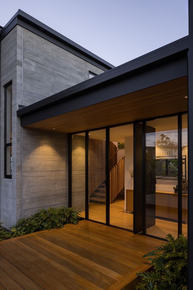 A contemporary multi-level residential entrance viewed from a timber deck at dusk. The facade combines large-format grey concrete wall panels, black metal framing, expansive glazed doors, a deep dark roof overhang, and a warm timber-lined soffit. Interior stairs and timber flooring are visible through the glazing, with integrated planting along the deck edges.