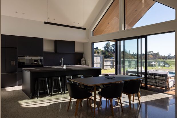 Contemporary open-plan kitchen and dining area with a large matte-black island, integrated dark cabinetry, built-in appliances, and four bar stools. A timber dining table with dark upholstered chairs sits beside expansive black-framed glazing and sliding doors, opening to a deck and landscaped outdoor setting. The room has a high angled ceiling with timber-lined sections, polished terrazzo-style flooring, and abundant natural light.