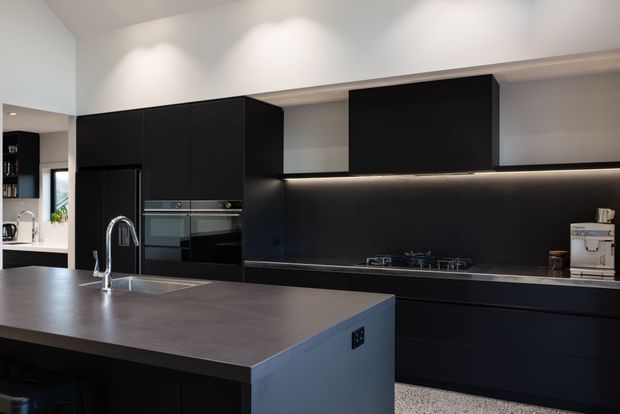 A contemporary kitchen with a large island, integrated sink and curved chrome tap, matte black full-height cabinetry, black splashback and dark benchtops. Built-in ovens and a coffee machine are integrated into the cabinetry, while recessed ceiling spotlights and concealed under-cabinet lighting illuminate the space. A small adjoining area is visible at left.