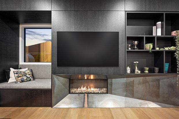 A dark, contemporary living room feature wall with a large wall-mounted television above a built-in linear gas fireplace. Black and charcoal timber-look cabinetry and open shelving frame the fireplace and display objects, while a grey upholstered window seat occupies the left side. Warm timber flooring contrasts with the dark joinery, and a small window provides a view outside.