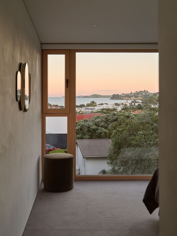 A narrow, softly lit bedroom or landing with a full-height timber-framed window overlooking a coastal residential landscape and water at sunset. The room has pale textured walls, a grey carpeted floor, a small round upholstered stool, and two round wall mirrors. A partial bed edge is visible at the right, confirming the space as a bedroom. The atmosphere is minimal, calm, and refined, with the expansive harbour view as the main feature.