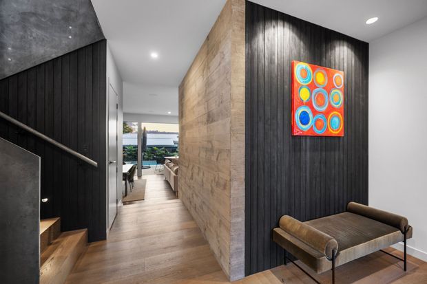 A contemporary residential hallway and entry landing viewed from near a staircase. The space features dark vertical timber wall panelling, a textured light stone or concrete feature wall, warm timber flooring, recessed ceiling lighting, and a compact leather bench beneath a colourful abstract artwork. A living area and exterior outlook are visible at the far end.