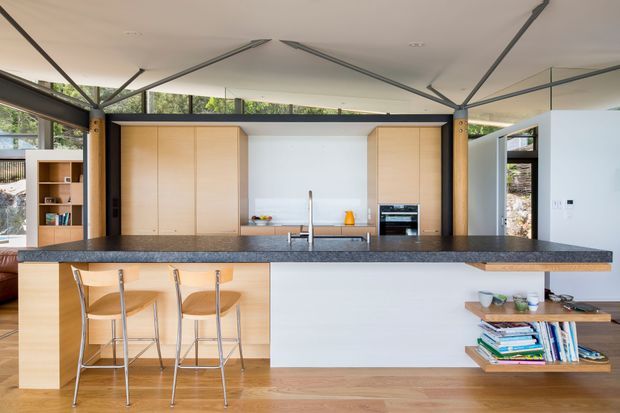A bright, open-plan contemporary kitchen with a large dark stone island and integrated breakfast bar seating. Light timber cabinetry frames the rear cooking zone, with a stainless-steel sink and tap, built-in oven, and open shelving. Exposed dark structural posts and beams, extensive glazing, and a white geometric ceiling create a spacious, modern interior. A low timber display shelf with books and decor sits at the right.