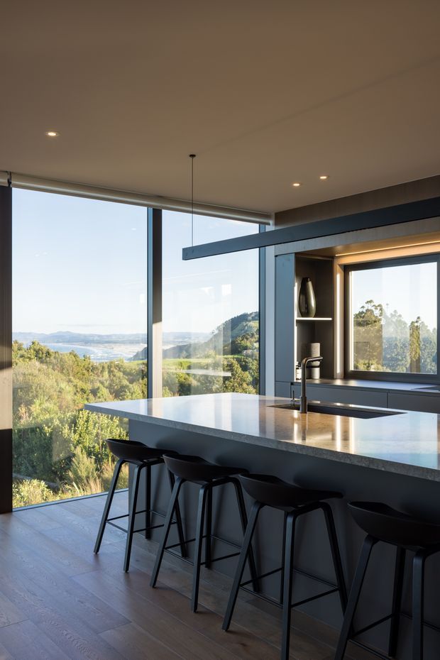 A refined contemporary kitchen with a large pale stone island and integrated sink, lined with black bar stools. Floor-to-ceiling glazing provides expansive views over a wooded, hilly landscape, while dark window frames, recessed lighting, and a sculptural linear pendant create a sleek, modern interior.