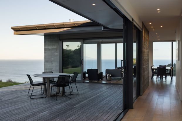 A contemporary coastal home interior opening onto a large timber deck overlooking the ocean. The image shows a glazed living area with dark-framed sliding doors, concrete walls, timber flooring, recessed ceiling lights, and a dining area extending down a long hallway.