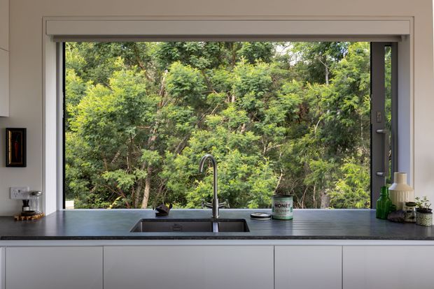 A contemporary kitchen sink bench positioned beneath a wide operable window, with a dark grey benchtop, integrated black sink, tall chrome tap, and white handleless cabinetry. The window frames dense green bushland, creating a strong indoor–outdoor connection. Small countertop accessories and artwork are visible, but the focus is the kitchen work surface and glazing.