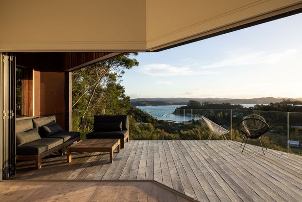 Open-plan indoor living area extending onto a large timber deck, with a dark upholstered sectional sofa, coffee table, and lounge chairs beneath a broad projecting roof. Full-height openings and glass balustrading frame a panoramic coastal or waterfront landscape.