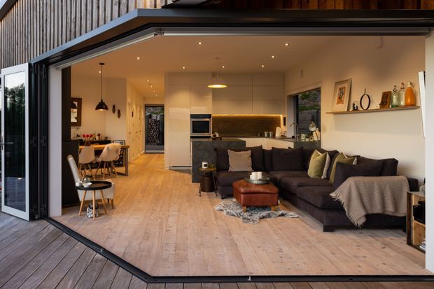 Open-plan contemporary living space viewed from a timber deck through fully opened folding glass doors. The room combines a dark sectional sofa and coffee table with a compact kitchen featuring pale cabinetry, a dark benchtop and integrated appliances. A dining area sits toward the rear, while timber flooring continues across the interior and deck, creating a seamless indoor-outdoor connection.