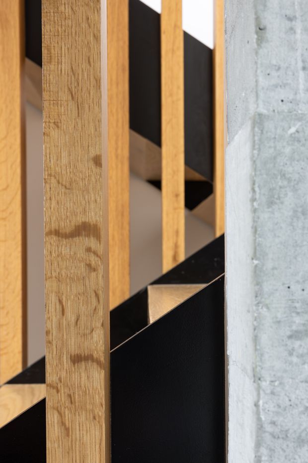 Close-up architectural detail of a stairway featuring warm timber vertical balusters and treads, contrasting black metal stair panels and balustrade elements, and a raw exposed concrete wall. The composition highlights material contrast and geometric lines rather than a complete room.