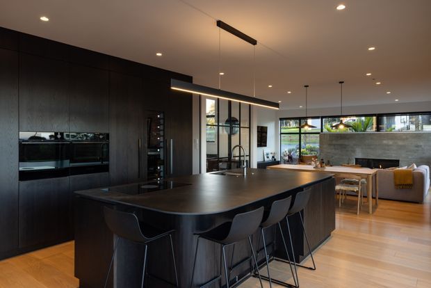 A sophisticated open-plan kitchen and dining area dominated by a large dark island with integrated seating for several people. Full-height dark timber cabinetry incorporates built-in appliances along the left wall, while a long linear pendant light hangs above the island. The space opens to a dining and living area with light timber flooring, extensive rear glazing, and a textured concrete feature wall. The palette is dark and refined in the kitchen, balanced by warm timber flooring and natural daylight.