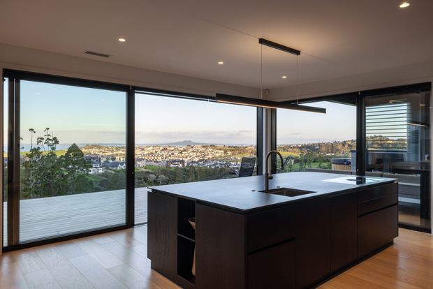 Contemporary kitchen interior with a large dark island, integrated sink and black tap, extensive black-framed floor-to-ceiling glazing, and a timber-toned floor. The open glazing overlooks a suburban coastal landscape and a broad deck, while recessed ceiling lights and a linear pendant provide illumination.