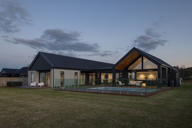 Exterior dusk view of a contemporary single-storey gable-roof residence with dark metal roofing and cladding, pale masonry walls, extensive glazing, and a warmly lit double-height gabled living area. A swimming pool and paved pool terrace sit directly in front of the house, with broad lawn surrounding the property.