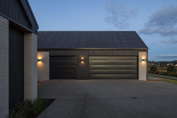 Twilight exterior view of a contemporary detached garage or outbuilding with two dark sectional garage doors, charcoal vertical cladding, pale masonry side walls, a low pitched metal roof, and warm wall-mounted lighting. A broad asphalt driveway occupies the foreground, with neighbouring residential landscape visible in the distance.