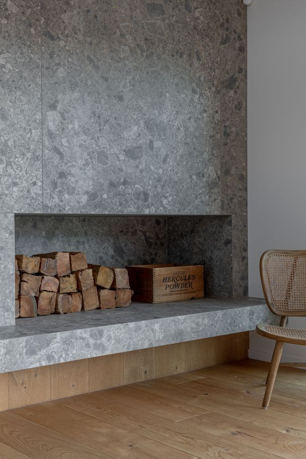 Close-up interior view of a sculptural fireplace clad in mottled grey natural stone, with an open firewood niche and wide stone hearth. Warm timber cabinetry and oak flooring contrast with the stone, while a woven cane chair sits at the right edge.
