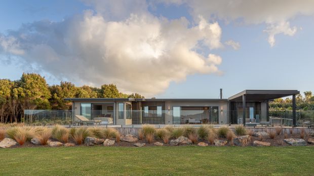 A single-storey contemporary home viewed from the landscaped front garden. The long, low-profile building has dark horizontal cladding, extensive floor-to-ceiling glazing, a flat roof, and a projecting covered outdoor area. Native-style grasses, rocks, and mature planting frame the facade.