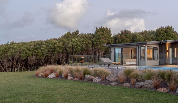 A low, single-storey contemporary home or pavilion set within a landscaped rural or bush-edge site. The dark flat-roofed building has extensive glazing, timber-toned wall cladding, a covered outdoor terrace, and a long swimming pool along the facade. Ornamental grasses, boulders, lawn, and dense trees surround the building under a cloudy sky.