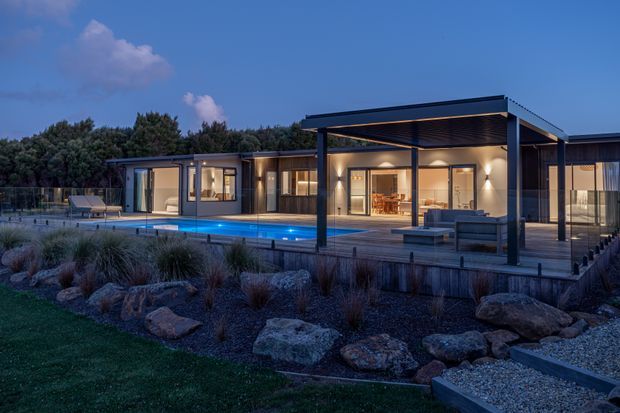 Contemporary single-storey residence at dusk, viewed from a landscaped garden. The home features extensive glazed openings, a flat-roofed pavilion, a covered outdoor entertaining deck, and a long swimming pool with low-maintenance rock and ornamental-grass landscaping.