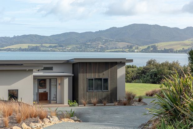 A contemporary single-storey residence with a low, flat roofline, smooth light-coloured walls, and a dark vertical timber-clad feature volume. The house is set within a landscaped driveway and garden overlooking a calm body of water and hilly rural terrain.