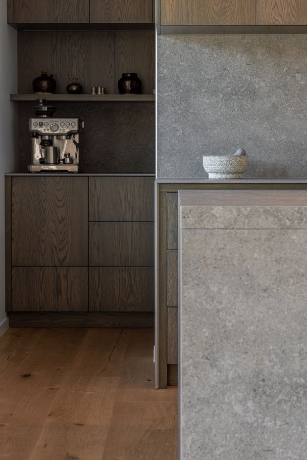 Close-up interior view of a refined kitchen with dark wood-grain cabinetry, a built-in coffee station, and a large grey stone island or benchtop in the foreground. The space has warm timber flooring, integrated joinery, open shelving with ceramic vessels, and a muted, sophisticated material palette.