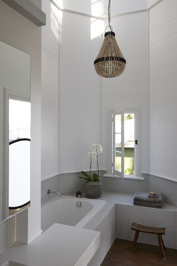 Bright, tall bathroom with white shiplap-style wall panelling, a deep freestanding bathtub set into grey mosaic-tiled surrounds, and a matching tiled vanity or ledge. A large woven pendant light hangs above the bath, while a small white-framed window, orchid, folded towels, and rustic wooden stool add warmth. A mirror is visible on the left wall.