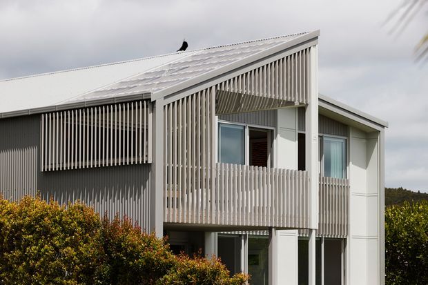 Exterior view of a contemporary multi-level residence with a sharply pitched roof, pale metal cladding, and extensive vertical timber screening. A sheltered upper-level balcony is enclosed by vertical slats, with large windows and glazed doors beneath a projecting roof canopy. Dense shrubs partially obscure the lower facade, and a wooded landscape is visible in the distance under an overcast sky.