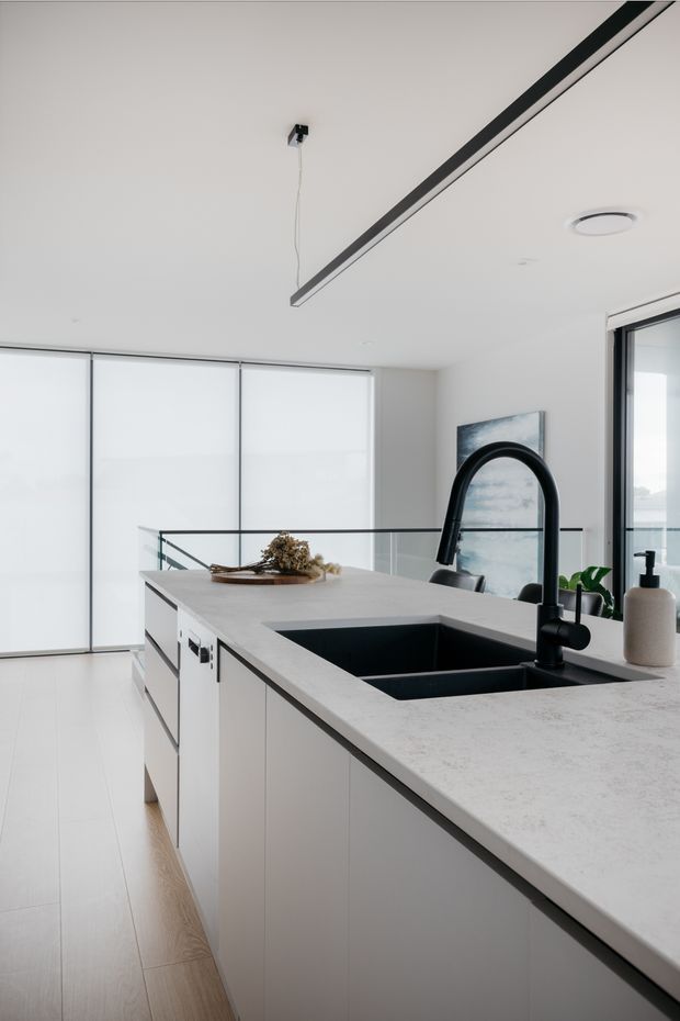 Bright contemporary kitchen interior dominated by a large pale stone island with integrated black double sink and matte-black gooseneck tap. White flat-panel cabinetry, light timber flooring, full-height translucent glazing, glass balustrade, recessed lighting and a linear black ceiling fixture create a clean, minimalist aesthetic.