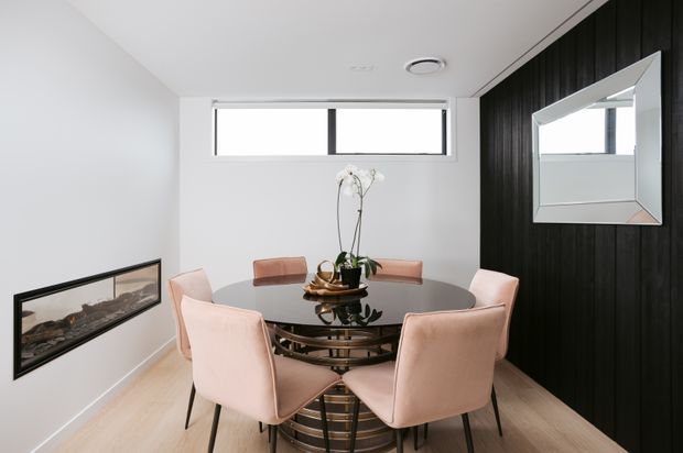 A compact, refined dining room with a round glass dining table and six blush upholstered chairs. The room features pale timber flooring, white walls, a long horizontal built-in fireplace, and a dramatic black vertical timber-panelled feature wall with a large frameless mirror. High horizontal windows provide natural light.