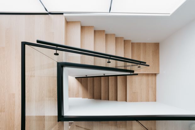 A minimalist interior stairwell featuring a sculptural timber-clad staircase or stair enclosure with repeated vertical wood panels and black-framed glass balustrades. Linear black track lighting runs along the ceiling and stair edge, while a large white ceiling plane and pale timber surfaces create a bright, clean, contemporary composition.