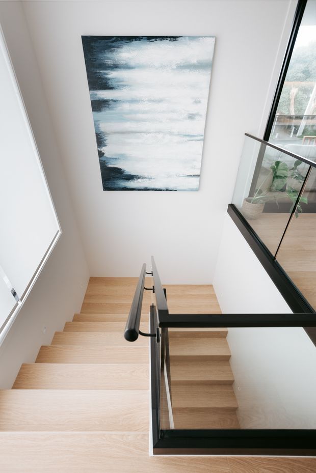 Contemporary interior view looking down a bright stairwell with pale timber stairs, white walls, and a black metal-framed glass balustrade. A large abstract blue-and-white artwork hangs on the stairwell wall, while an upper-level landing with a potted plant is visible through the glazing.