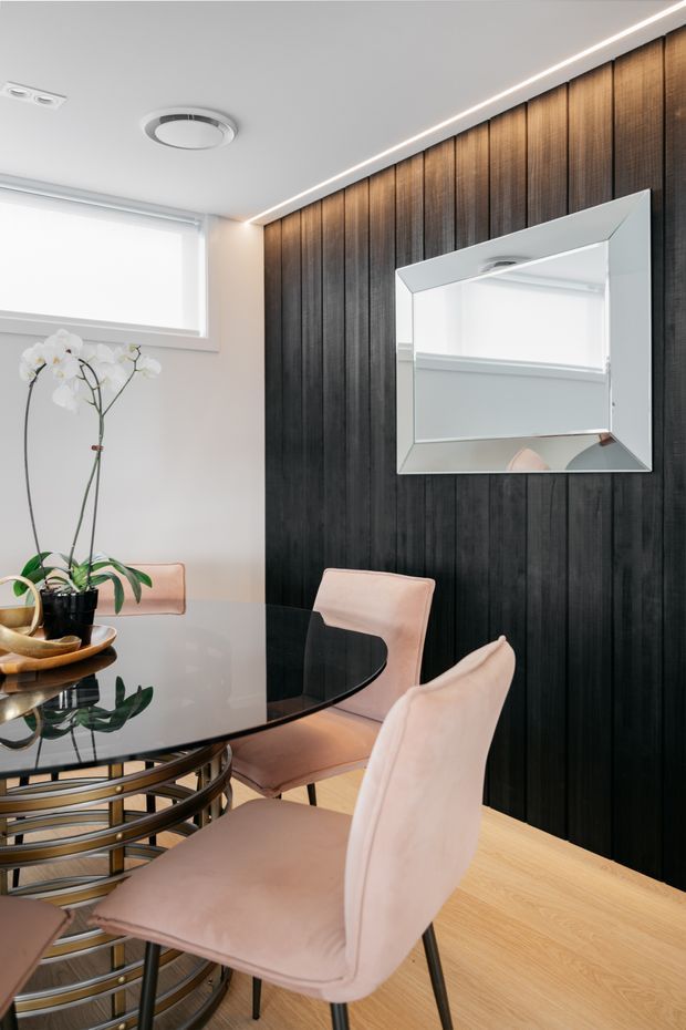 A refined dining area with a round black glass dining table and upholstered blush-pink chairs. The room features a dramatic full-height dark timber vertical-panel feature wall with a large geometric framed mirror, pale walls, light timber flooring, recessed ceiling lighting, and a white orchid as a restrained decorative element.