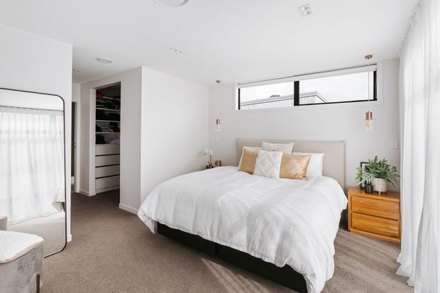 Bright contemporary bedroom with a neatly made bed, soft white bedding, a slim horizontal clerestory window, sheer full-height curtains, wall-mounted bedside lights, a timber chest of drawers, and an open walk-in wardrobe. Neutral finishes and abundant daylight create a calm, airy atmosphere.