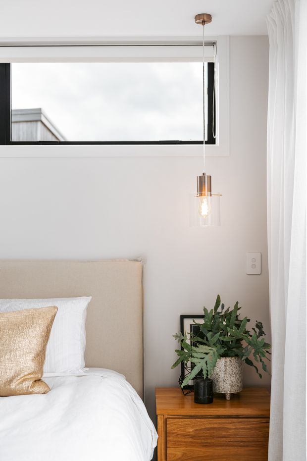 Bright, compact bedroom corner featuring a low upholstered beige headboard, crisp white bedding, a warm timber bedside cabinet, potted greenery, a framed artwork, sheer white curtains, and a suspended glass-and-metal pendant light. A long horizontal window provides natural light, creating a calm, contemporary interior.