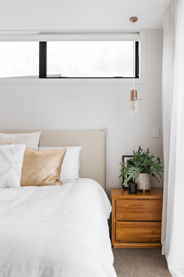 Bright, softly styled bedroom featuring a low upholstered beige bedhead, layered white and textured neutral bedding, a warm timber bedside table, indoor greenery, sheer white curtains, and a black-framed horizontal window. A minimalist pendant light hangs beside the bed.